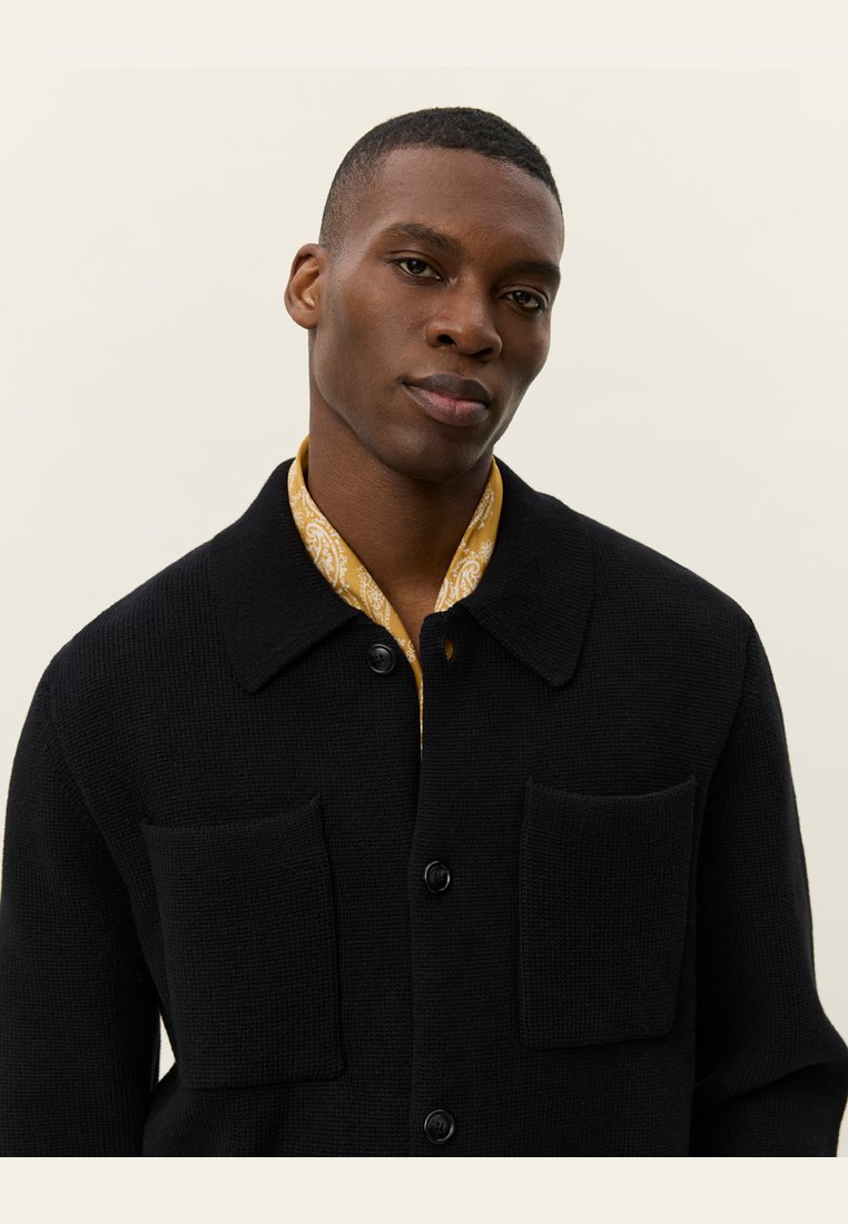 Schwarze Strickjacke mit zwei aufgesetzten Taschen, Knopfverschluss und Kragen, über einem gelben gemusterten Hemd getragen.