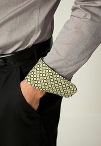 Cămașă de dress cu mânecă lungă în violet deschis, cu un model texturat, având un manșon cu un pattern contrastant verde și auriu. Curea neagră vizibilă.
