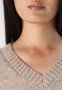 Pull en maille texturé beige avec décolleté en V, mettant en valeur des détails côtelés autour du col et une peau lisse visible au niveau du cou.