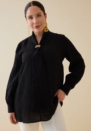 Kvinde med mørkt hår i en elegant knold iført sort bluse med bindedetalje foran og store guldringe, kombineret med hvide bukser mod en ensfarvet baggrund.