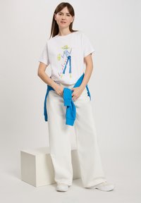 Biała koszulka z kolorową ilustracją postaci w stroju plażowym, zestawiona z luźnymi białymi spodniami i niebieskim swetrem związanym w talii.