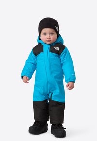 Lyseblå vinterdress med svarte detaljer, som har hette, elastiske mansjetter og glidelås foran; svarte vinterstøvler fullfører antrekket.