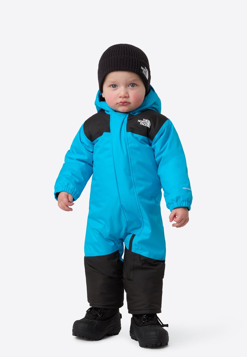 Lyseblå vinterdress med svarte detaljer, som har hette, elastiske mansjetter og glidelås foran; svarte vinterstøvler fullfører antrekket.