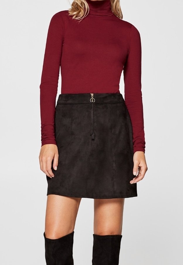 Femme portant un col roulé bordeaux à manches longues, une mini-jupe en suède noire avec une fermeture éclair devant et des bottes noires montant jusqu'aux genoux, debout devant un fond uni.