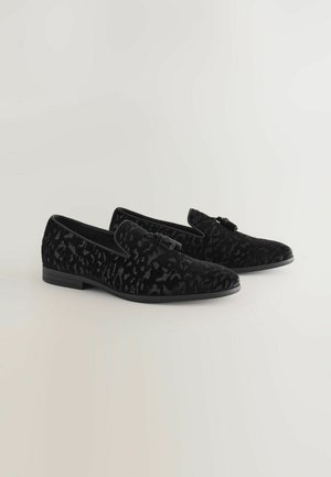 Schwarze Samtloafer mit strukturierter Oberfläche, ausgestattet mit einem dezenten Muster und einer kleinen Schleifenapplikation an der Seite. Flache Sohle, klassisches Design.