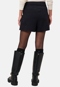 Shorts noirs ajustés avec une finition lisse, comportant deux poches arrière. Le modèle porte des bottes noires brillantes arrivant au genou avec des dessus en tricot noir doux.