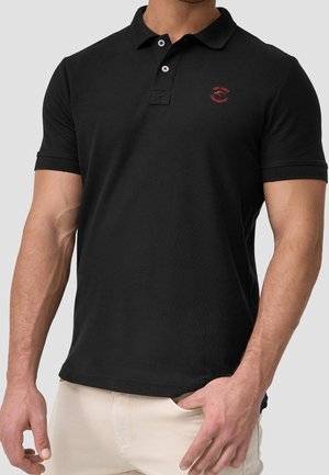 Uomo con polo nero a maniche corte con logo rosso sul petto, pantaloni beige e una mano in tasca su uno sfondo semplice.