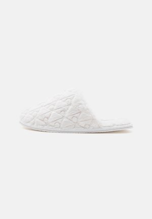 Zapatilla blanca acolchada con punta abierta, parte superior de tela suave y suela plana, mostrada de lado sobre un fondo liso.