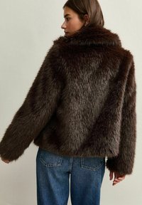 Donna che indossa una giacca in pelliccia sintetica marrone scuro e jeans blu, rivolta di lato con la testa girata a sinistra su uno sfondo neutro.