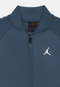 Jachetă bleumarin cu fermoar, textură striată, având un logo Jordan alb pe pieptul stâng și design cu guler înalt.