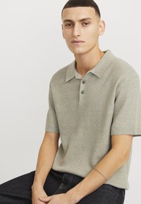 Polo de manga corta en un tejido de punto gris claro, con cuello, tapeta de dos botones y detalles sutiles, combinado con pantalones de mezclilla oscuros.
