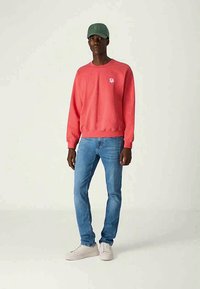 Man med grön keps, korallfärgad långärmad sweatshirt med liten blommig logotyp, blå jeans och vita sneakers som står mot en enfärgad bakgrund.