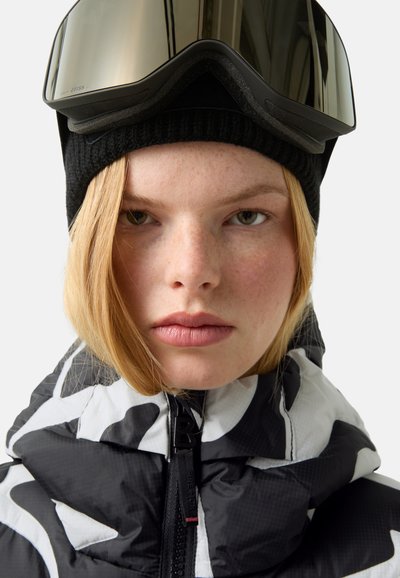 Černobíle vzorovaná nadýchaná bunda s vysokým límcem, doplněná černou pletenou čepicí a zrcadlovými lyžařskými brýlemi. Hladká textura.