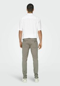 Camisa blanca de manga corta, pantalones grises con dos bolsillos traseros y zapatillas blancas, vistos desde atrás sobre un fondo claro.