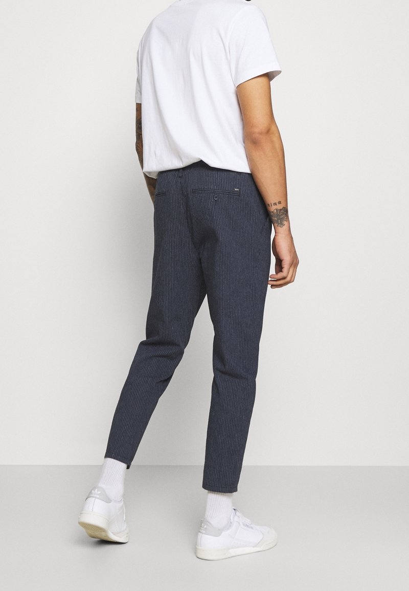 Pantalones de mezcla de algodón en azul marino con un patrón de rayas verticales, corte ajustado, dos bolsillos traseros y detalles en las costuras laterales. Llevado con una camisa blanca.