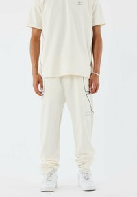 Camisa de manga corta de color crema claro con un pequeño logo, combinada con pantalones a juego que presentan detalles de ribetes laterales, ambos confeccionados en un material suave.