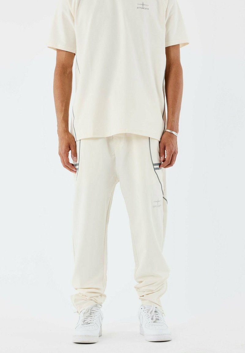 Camisa de manga corta de color crema claro con un pequeño logo, combinada con pantalones a juego que presentan detalles de ribetes laterales, ambos confeccionados en un material suave.