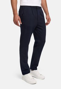 Férfi, aki tengerészkék, keskeny szabású nadrágot visel oldalzsebekkel és fehér sportcipővel, egyszerű háttér előtt áll.