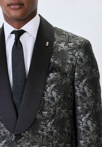 Black floral patterned tuxedo jacket with satin lapels, paired with a black dotted tie and a pocket square. Gold skull pin detail on lapel.