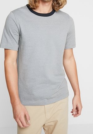 Uomo che indossa una camicia a maniche corte grigia chiara con colletto nero abbinata a pantaloni beige, in piedi contro uno sfondo neutro.