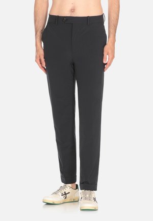 Pantaloni neri slim-fit con una texture liscia, tasche frontali e una piccola chiusura a bottone. Abbinati a sneakers beige e verdi.