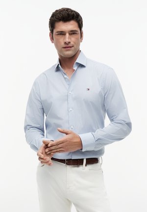 Homme portant une chemise à carreaux bleu clair à boutons avec le logo Tommy Hilfiger, pantalon blanc et ceinture marron, ajustant ses mains.