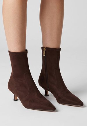 Stivaletti marroni in suede con punta appuntita, tacco basso kitten e zip laterali, indossati su gambe nude su sfondo semplice.