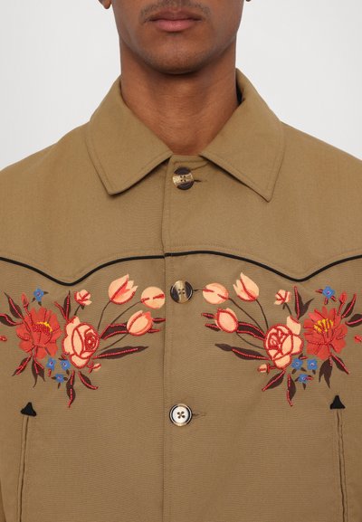 Tan button-up jacket featuring floral embroidery in red and blue, with black accents and a collared neckline. Buttons display a shiny finish.