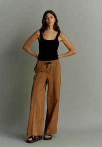 Top din bumbac negru cu falduri, asortat cu pantaloni largi maro, având cusături pe față și un cordon la talie. Modelul poartă sandale cu platformă.