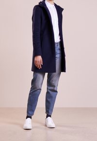 Navy wool coat with a structured design, featuring a high collar and button details, worn over a white shirt and blue jeans with white sneakers.
