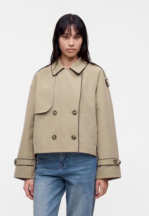 Femme portant une veste courte beige à double boutonnage avec un bord noir et un jean bleu, debout sur fond blanc.