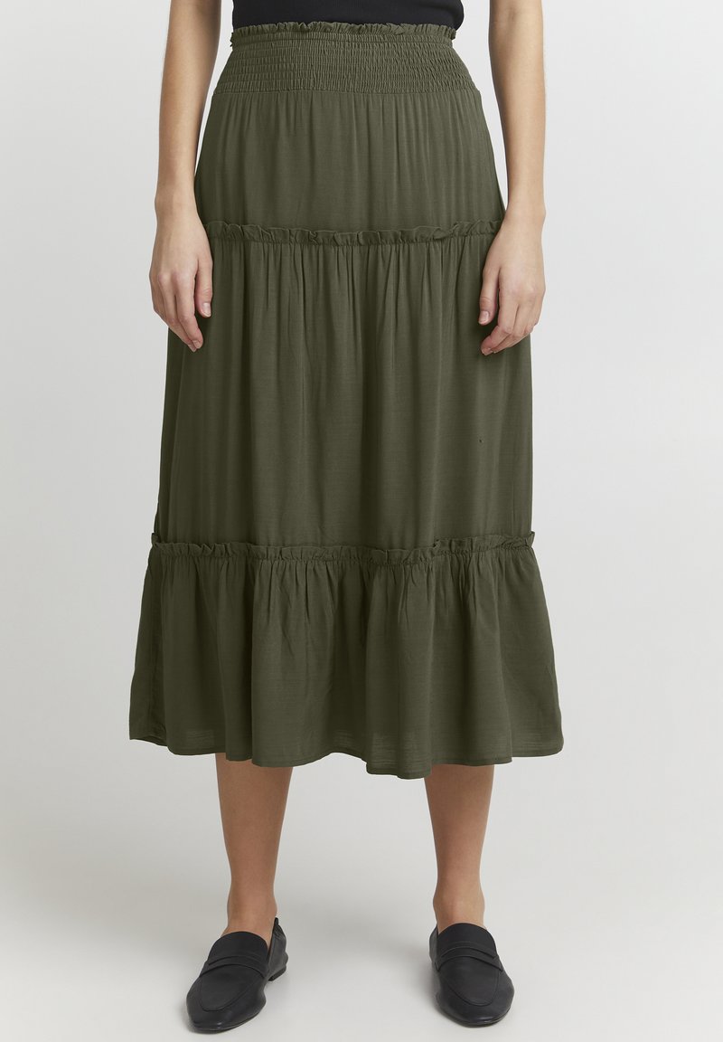 Gonna lunga a strati di colore verde oliva con vita smock, tessuto fluido e orlo arricciato. Indossata con scarpe scivolate scure.