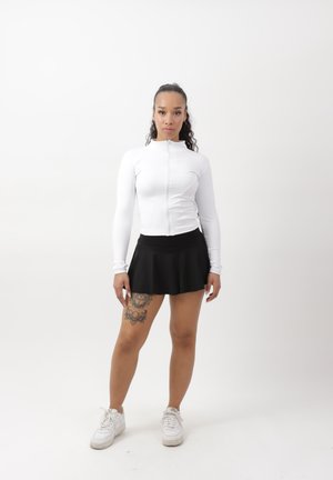 Frau steht, trägt ein weißes Zip-up langärmliges Oberteil, schwarzen Rock, weiße Turnschuhe und ein florales Tattoo am linken Oberschenkel.