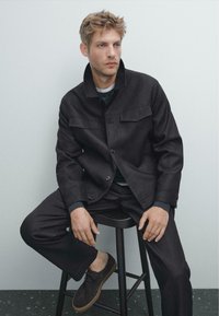 Svart utility-stil jacka med två bröstfickor, parat med matchande byxor och bruna skor, sittande på en pall mot en neutral bakgrund.