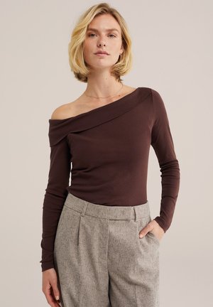 Lyshåret kvinde iført en mørkebrun off-shoulder langærmet top og lysegrå højtaljede bukser med hånden i lommen.