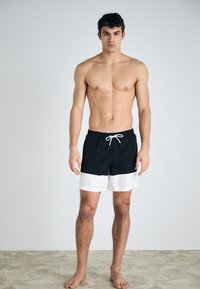 Svart og hvit svømmebadeshorts med elastisk midje og snøring, med en fargeblokkdesign. Modellen står barbeint på en strukturert overflate.