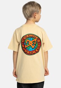 Beige t-shirt met korte mouwen met een kleurrafiek cirkelvormig ontwerp op de achterkant, waarop een wereldbol en gestileerde tekst in rood en groen zijn weergegeven.
