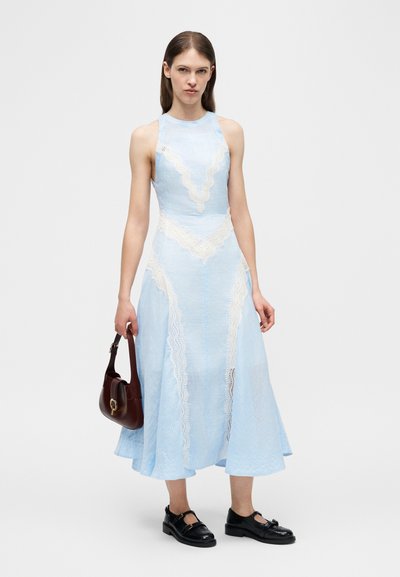 Νεαρή γυναίκα στέκεται φορώντας αμάνικο γαλάζιο μίντι φόρεμα με λευκές δαντελένιες λεπτομέρειες, κρατώντας σκούρο καφέ τσάντα και φορόντας μαύρα παπούτσια.