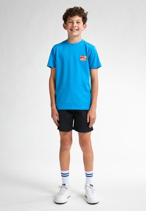 Camiseta de algodón azul brillante con mangas cortas, que presenta un pequeño gráfico rojo en el pecho. Combinada con pantalones cortos negros y zapatillas blancas.