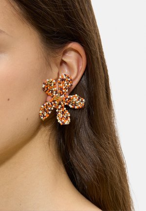 Zbliżenie ucha z dużą kolczykiem w kształcie kwiatu wykonanym z małych pomarańczowych, brązowych, białych i żółtych koralików.