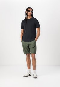 Schwarzes T-Shirt mit kurzen Ärmeln, grüne Shorts, Sonnenbrille, weiße Socken und Turnschuhe mit schwarz-grauen Akzenten, stehend vor einem weißen Hintergrund.