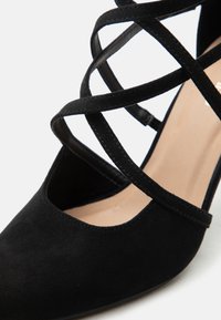 Γυναικεία μαύρα σουέντ παπούτσια με μυτερή μύτη, σταυρωτά λουριά στον αστράγαλο και μπεζ πάτο, φωτογραφημένα σε λευκό φόντο.