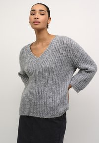 Pulover tricotat de un gri deschis, cu decolteu în V, mâneci 3/4 și o croială relaxată. Material texturat cu un aspect moale, asortat cu pantaloni închisi la culoare.