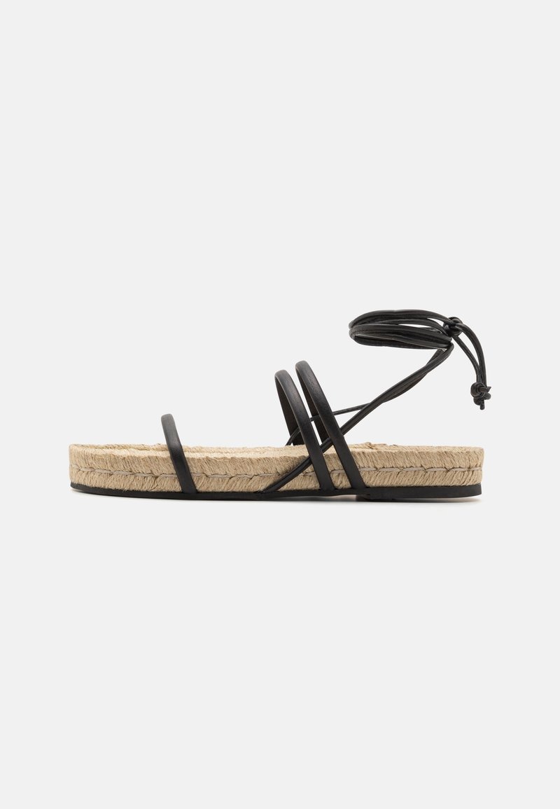 Sandale plate avec semelle plateforme en jute, fines lanières noires traversant le pied, et lien à nouer autour de la cheville sur fond gris clair.