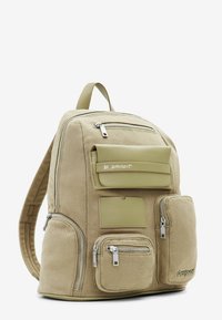 Béžový látkový batoh s několika kapsami na zip, klopnovou kapsou a vyraženým logem. Má texturovaný povrch a nastavitelné ramenní popruhy.