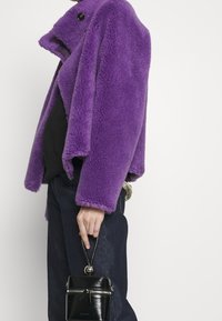 Livlig lila pälsjacka med en texturerad yta, rund krage och en enkel knappstängning, ihop med mörka jeans och en liten svart väska.