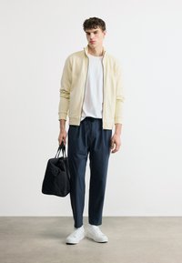 Νεαρός άνδρας που φοράει μπεζ ζακέτα με φερμουάρ, λευκό μπλουζάκι, ναυτικό παντελόνι, λευκά αθλητικά παπούτσια, κρατώντας μαύρη τσάντα duffel, στέκεται σε γκρι πάτωμα.