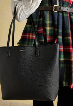 Schwarze Tote-Tasche aus strukturiertem Leder mit doppelten Griffen, gehalten von einer Person, die einen weißen Ärmel und ein kariertes Kleid mit braunem Gürtel trägt.