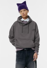 Szary hoodie z kieszenią kangurzą i sznurkami ściągającymi, nałożony na jasnofioletową koszulę w paski. Fioletowa dziana czapka z wydłużonymi uszami. Ciemne dżinsy.