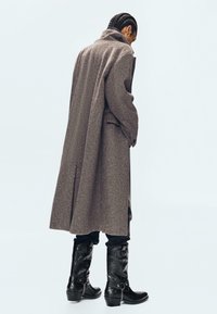 Dolga rjava volnena bunda z visokim ovratnikom, zadnjim razporkom in stranskimi žepi, v kombinaciji s črnimi usnjenimi škornji z dekorativnimi detajli.